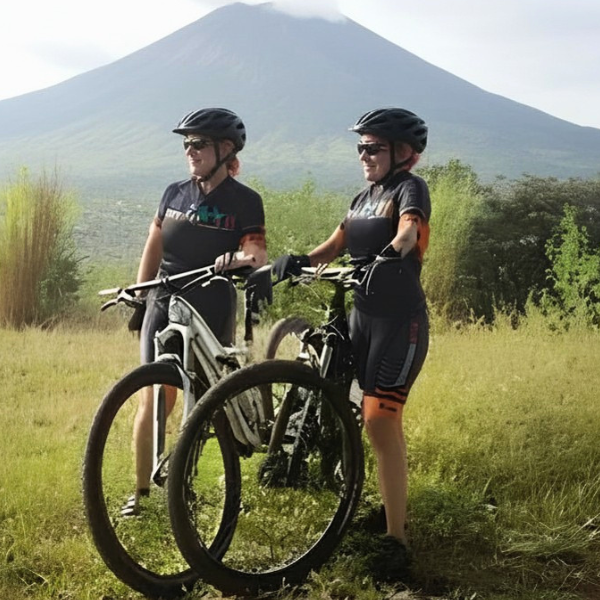 Two women with mountain bikes standing in a grassy field, wearing helmets and sunglasses, with a large volcano in the background.