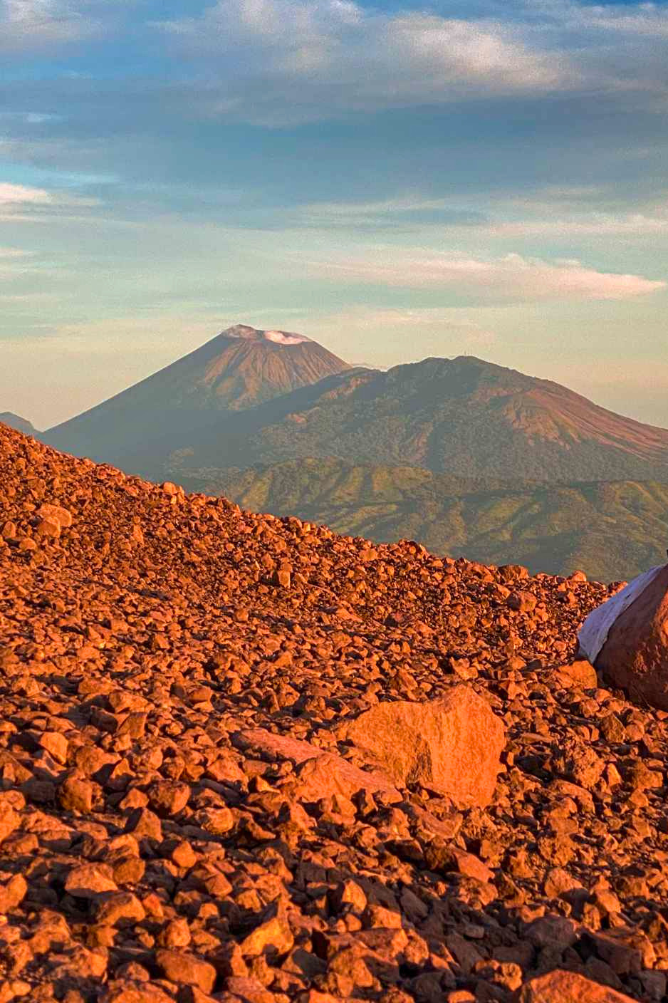 Volcanic landscape with reddish rocks in the foreground and a distant snow-capped volcanic mountain with a sky filled with soft clouds in the background.