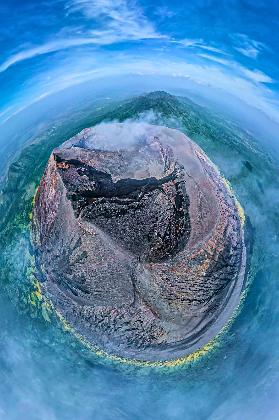 A distorted, wide-angle aerial view of a San Cristobal volcano with a summit crater, surrounded by blue ocean and distant landforms under a partly cloudy sky.