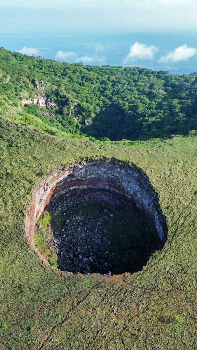 A large, deep crater in lush green landscape with rocky interior, surrounded by dense vegetation and rolling hills.