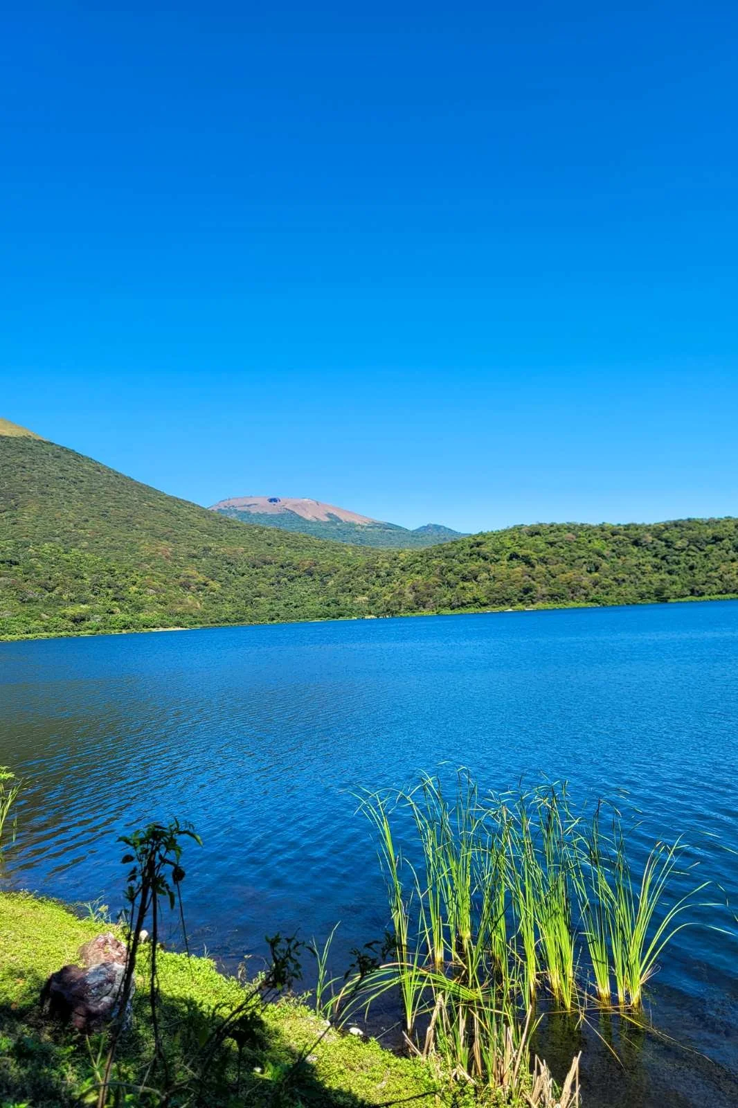 Scenic landscape with a blue lake, green hills, and mountains under a clear blue sky.