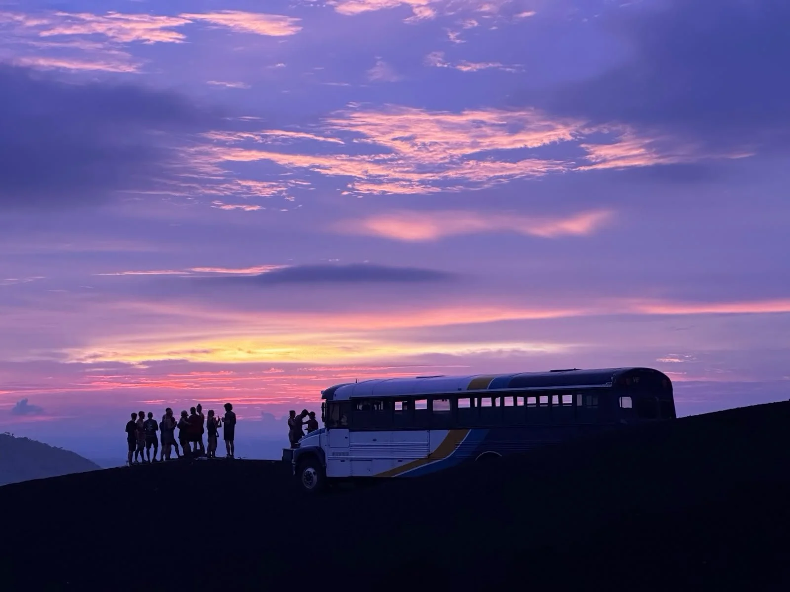 Silhouettes of people standing and gathering near a bus on a hilltop against a colorful sunset sky with pink, purple, and orange clouds.