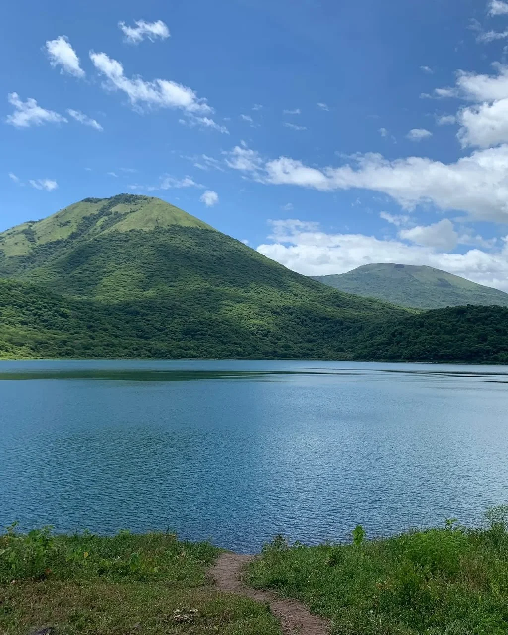 A peaceful lake with green hills and mountains in the background under a blue sky with white clouds.