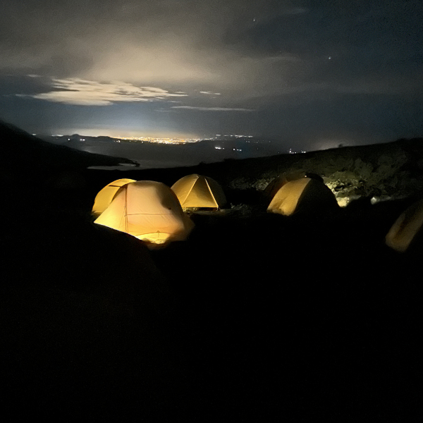 Nighttime camping scene with illuminated tents on a hilltop, dark sky with clouds and city lights in the distance.