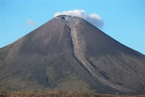 Momotombo volcano with a broad, conical shape and a small cloud of smoke or ash at its summit, against a blue sky.