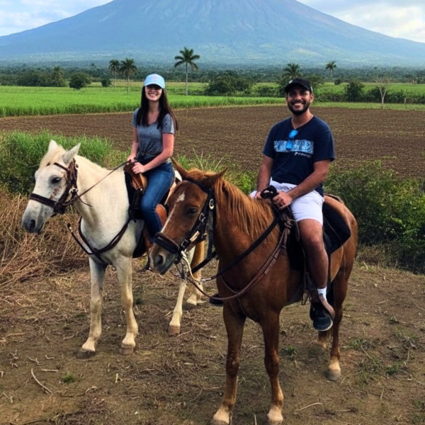 A man and woman riding horses in front of a volcano with lush greenery and palm trees.