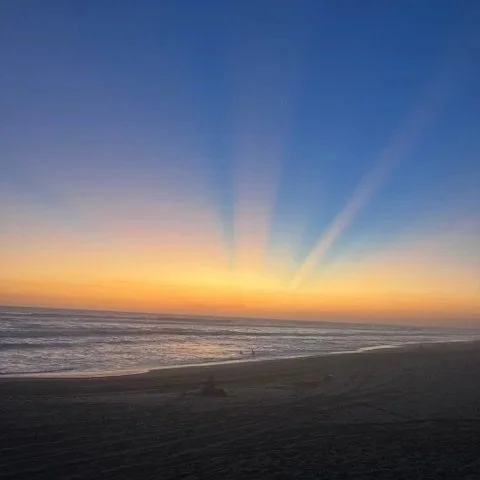 Sunset over the ocean with colorful rays of light in the sky and a sandy beach in the foreground.