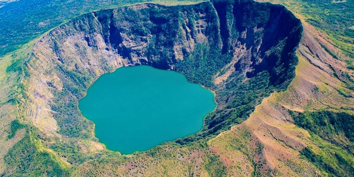 A volcanic crater filled with blue water, surrounded by steep, rocky slopes and green vegetation.