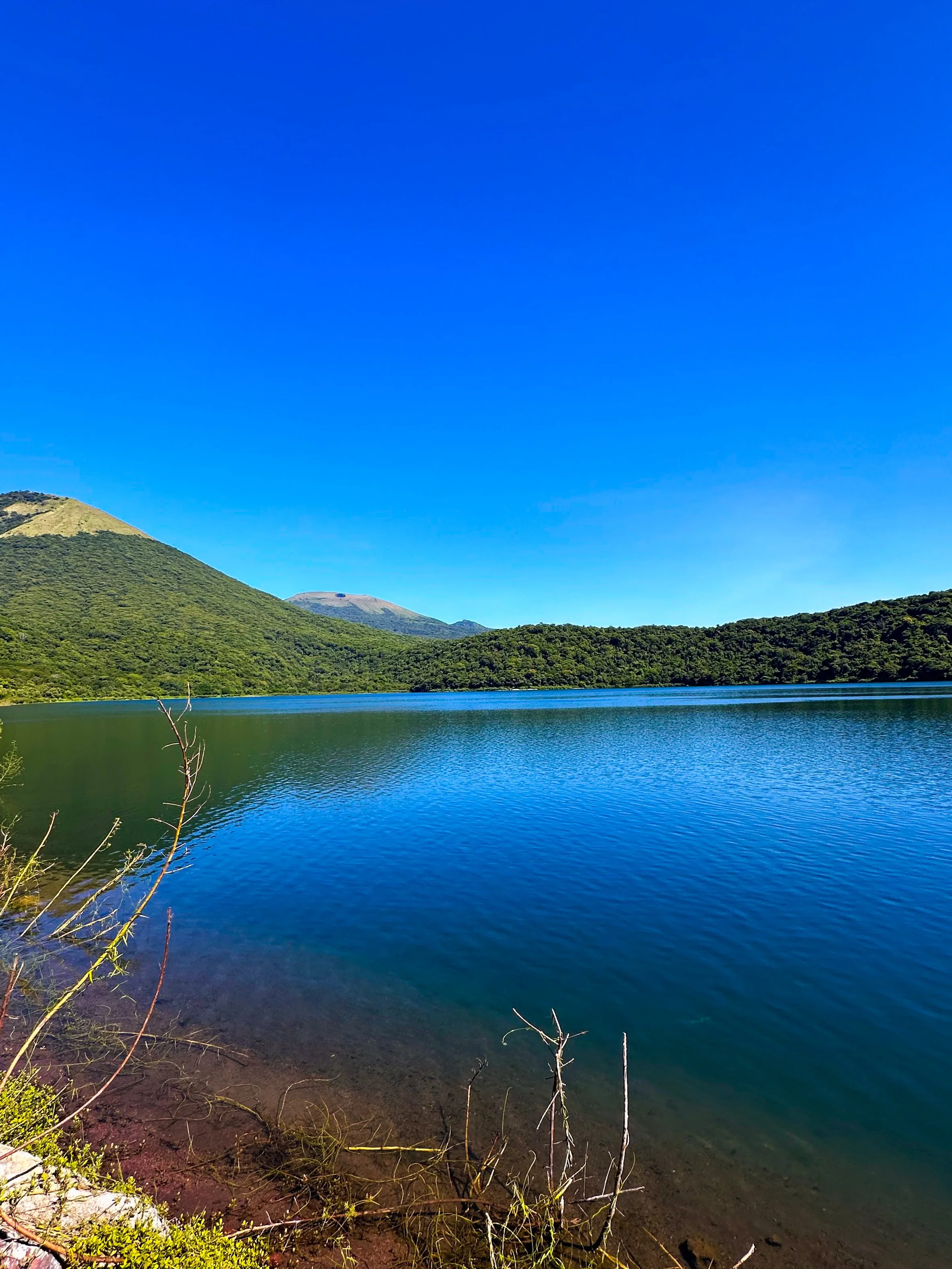 A scenic landscape featuring a calm lake surrounded by lush green hills and mountains, with a clear blue sky overhead.