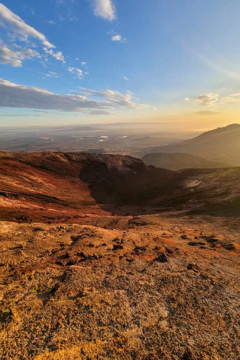 A volcanic crater with reddish-brown rocky terrain, overlooking a vast landscape under a partly cloudy sky during sunset or sunrise.