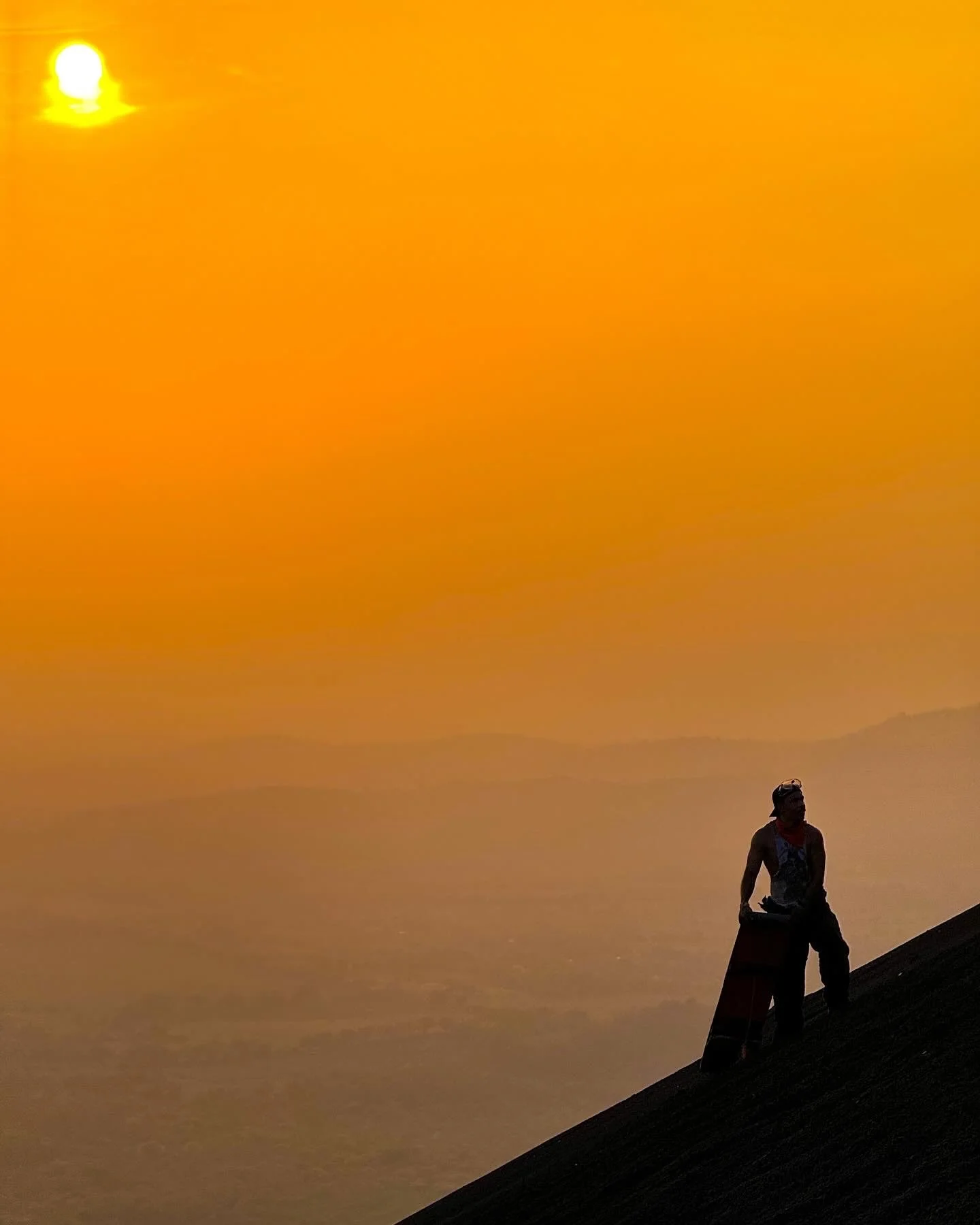 Person sitting on a slope at sunset with an orange sky and distant volcanoesin the background.