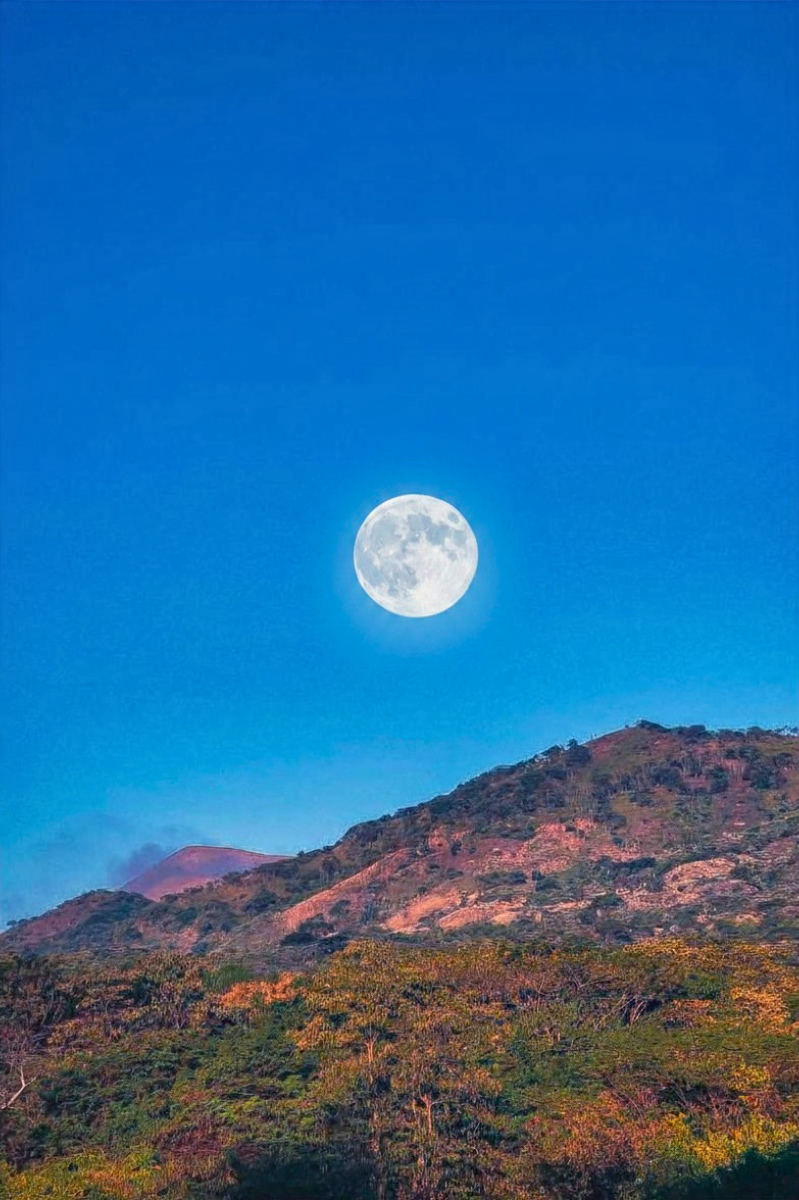 A full moon rising above Telica volcano with trees and shrubs under a clear blue sky.