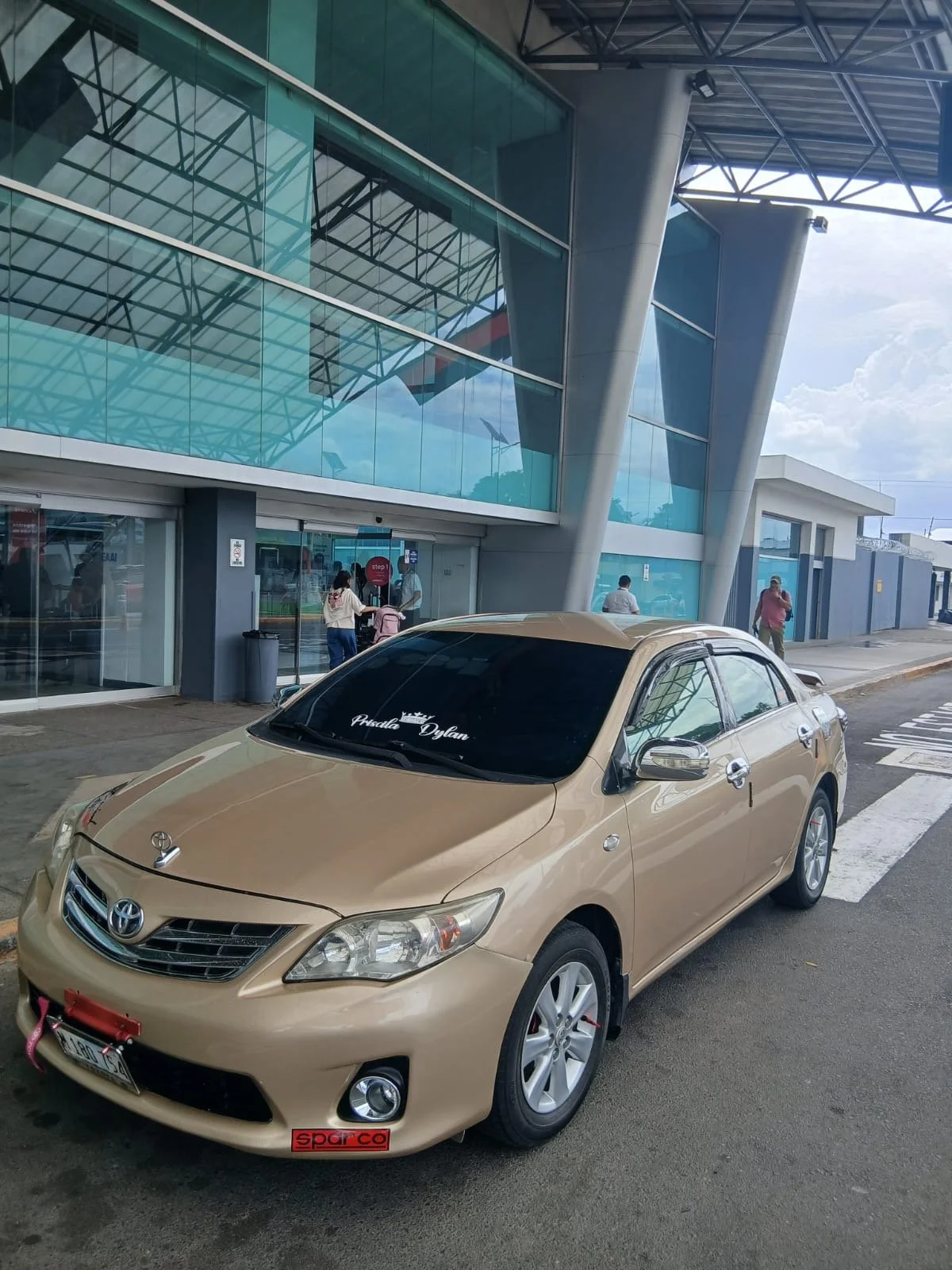 A beige Toyota sedan parked outside a modern glass building with people walking near the entrance.
