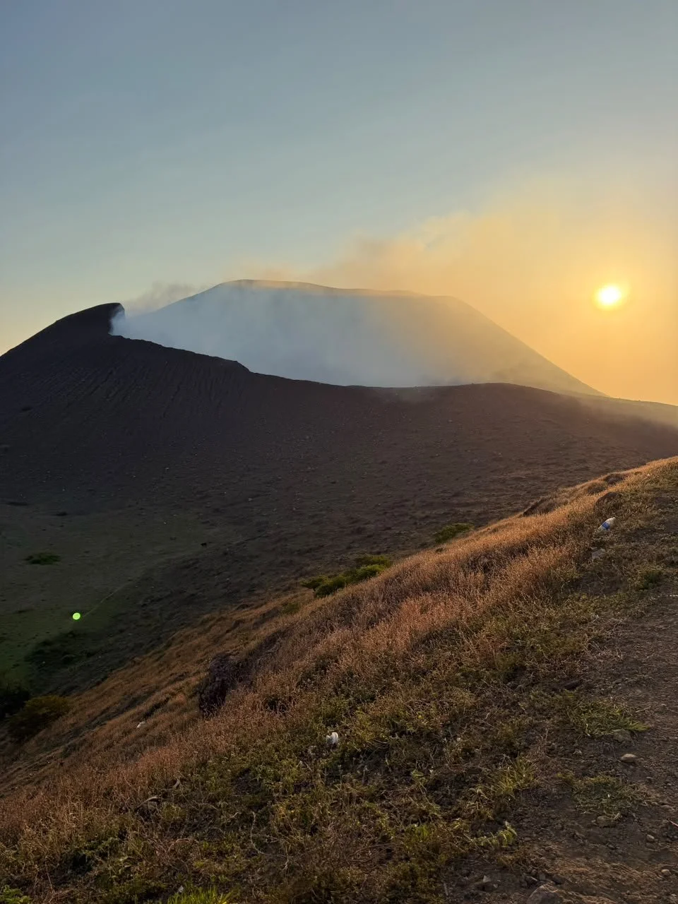 Sunset over a volcanic landscape with a smoking volcano, dark ash-covered slopes, and grassy foreground.