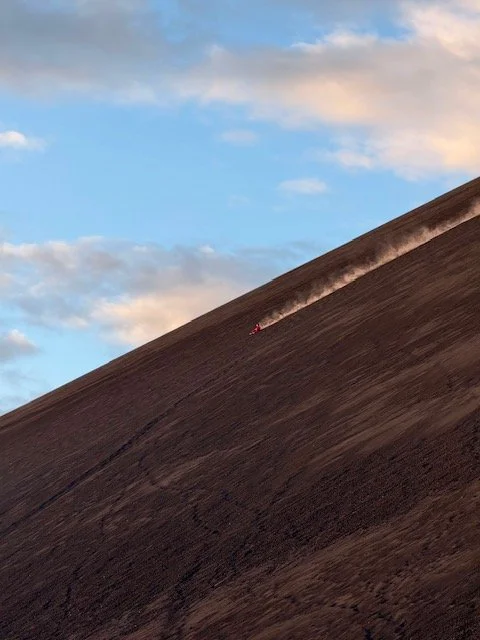 A small volcano boarder on a steep, dark hillside, leaving a trail of dust behind it, with a partly cloudy sky above.