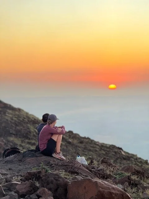 Two people, a woman and a man, sitting on a rocky hillside, watching a sunset over the volcanoes.