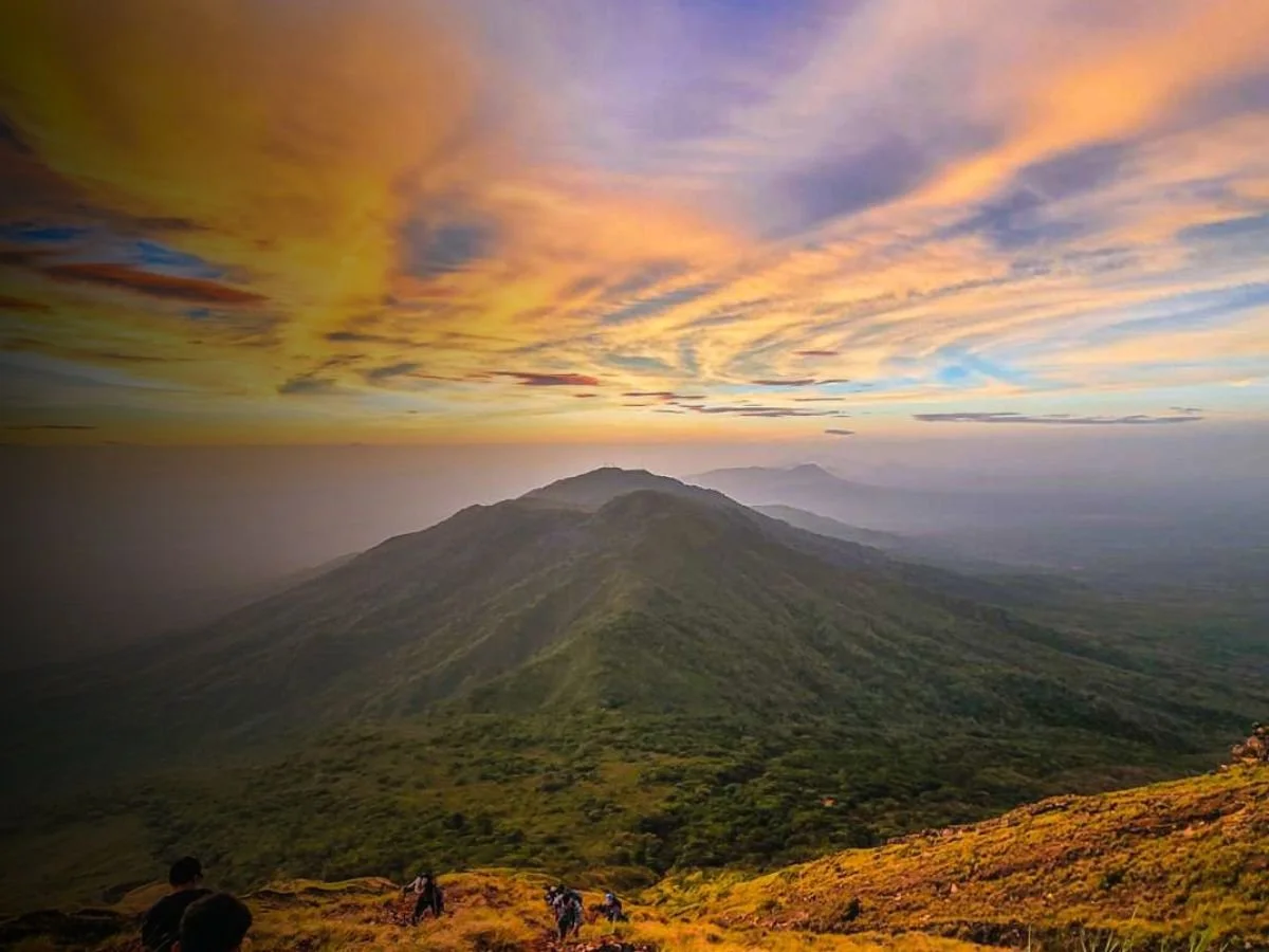 Sunset over a mountain landscape with vibrant clouds in the sky and hikers walking on a trail.