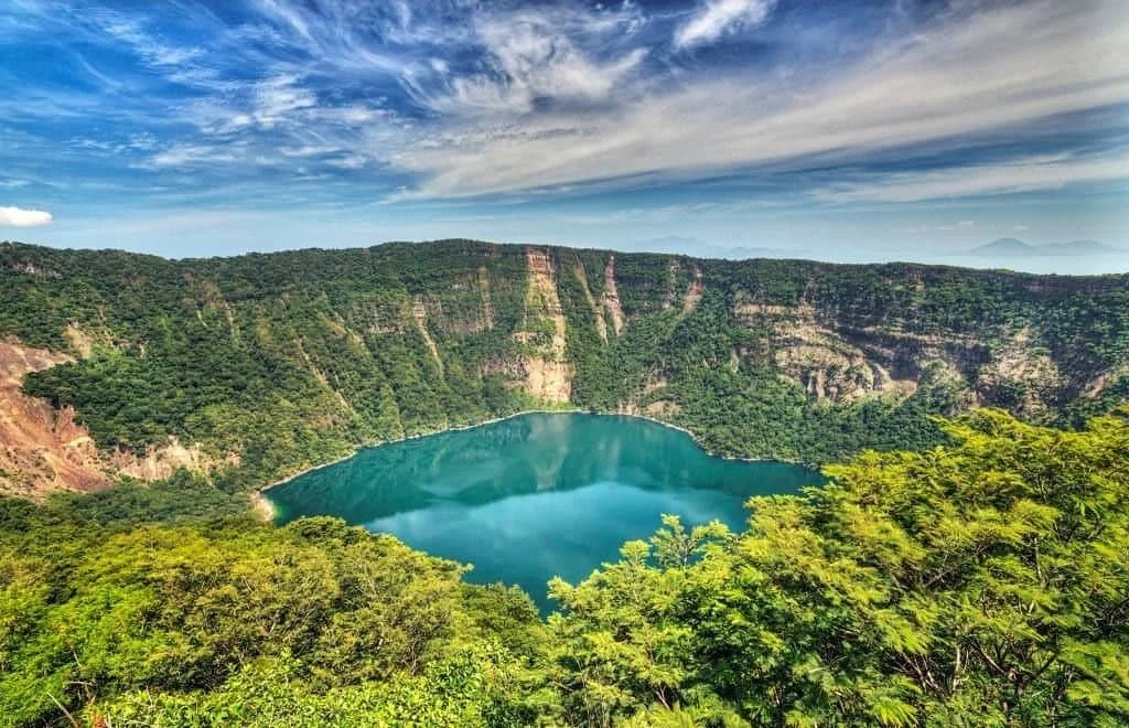A scenic view of Cosiguina volcanic crater lake surrounded by lush green forest and steep cliffs under a blue sky with wispy clouds.