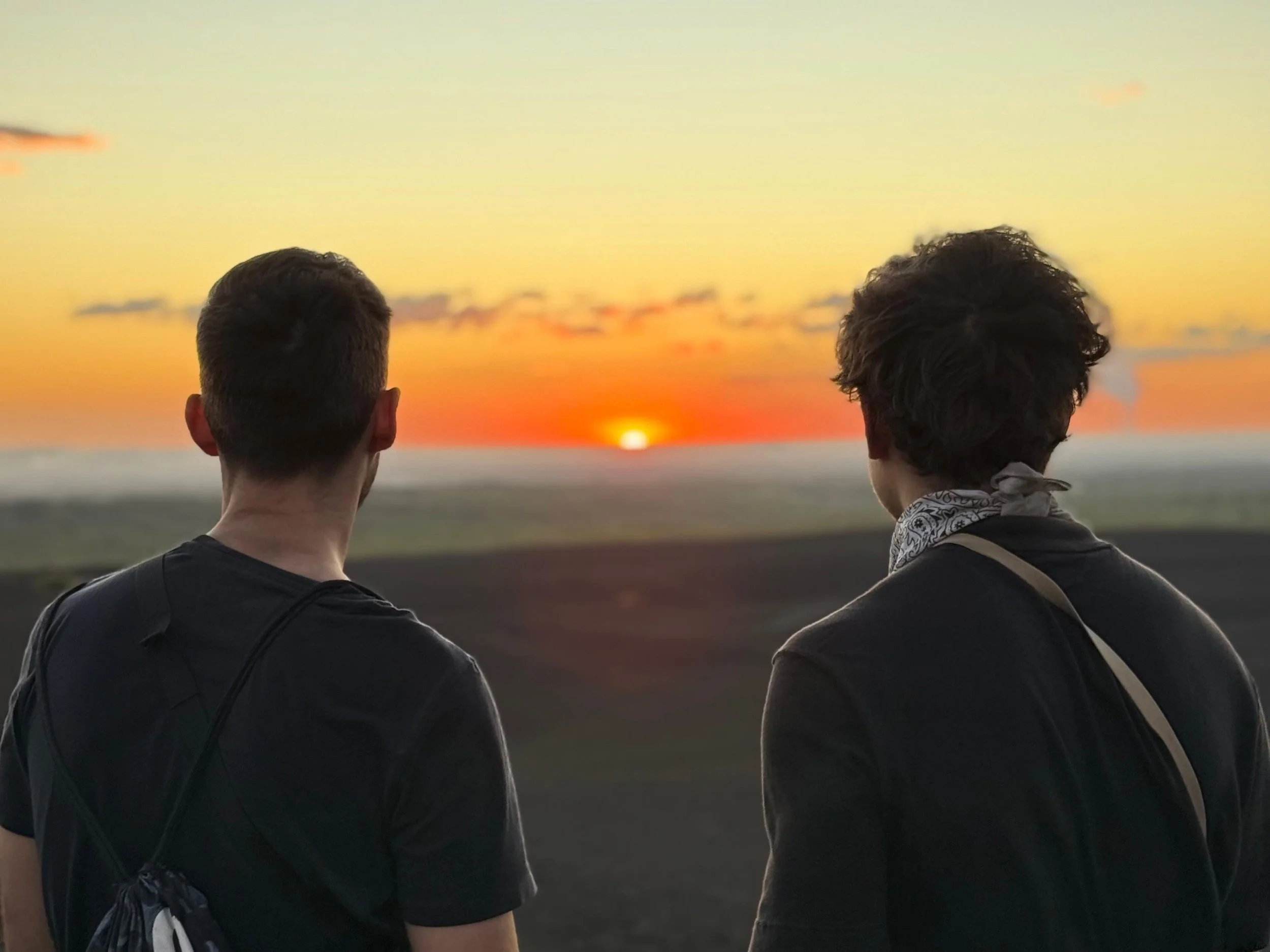 Two men with backpacks standing outdoors watching a sunset at dusk.