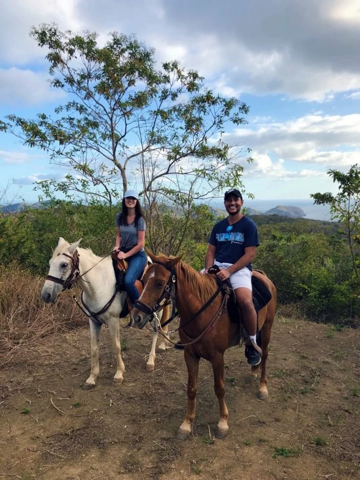 A man and woman riding horses on a scenic trail with green trees and mountains in the background.