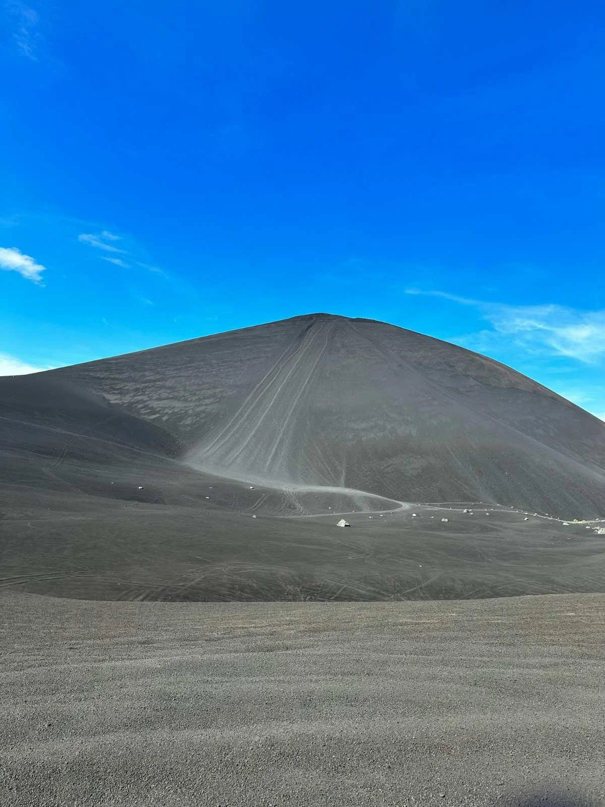 A cerro negro volcano with a grayish surface under a clear blue sky, with faint tracks and some scattered rocks on the ground in the foreground.