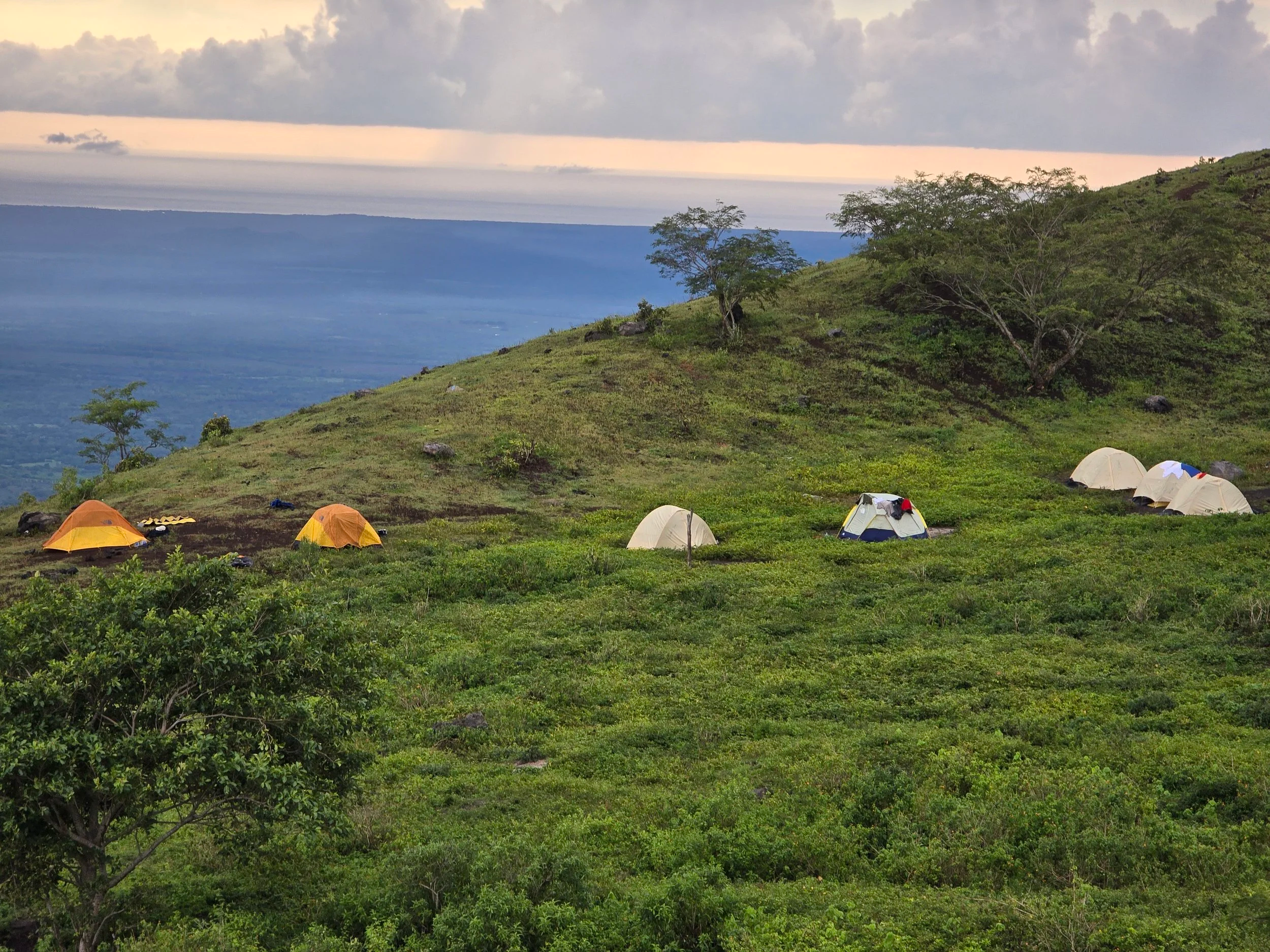 Tents set up on a green hillside overlooking a valley with mountains in the distance, cloudy sky overhead.