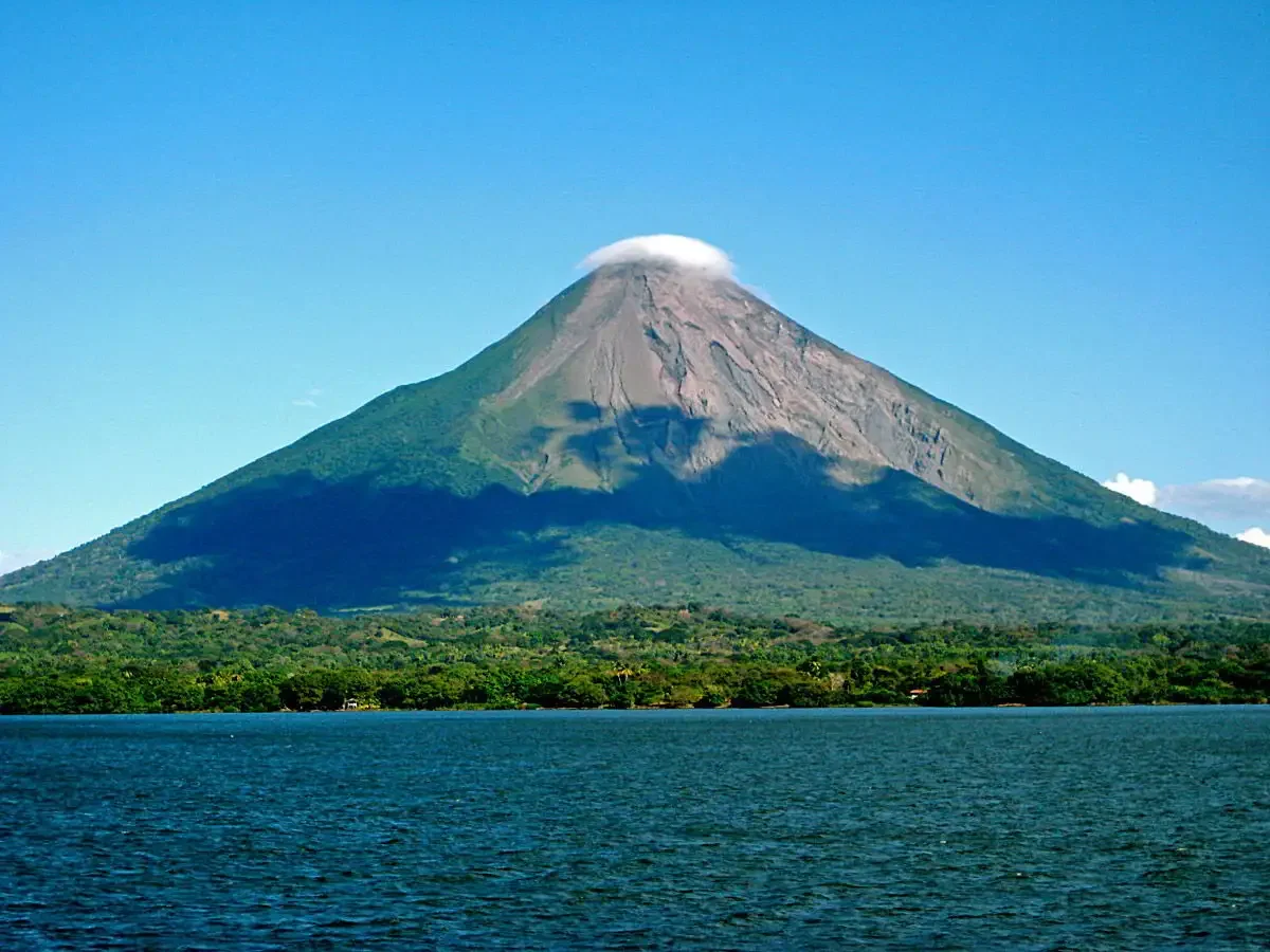 Momotombo volcano with a cloud at its crater, viewed from a body of water with green vegetation at the base.