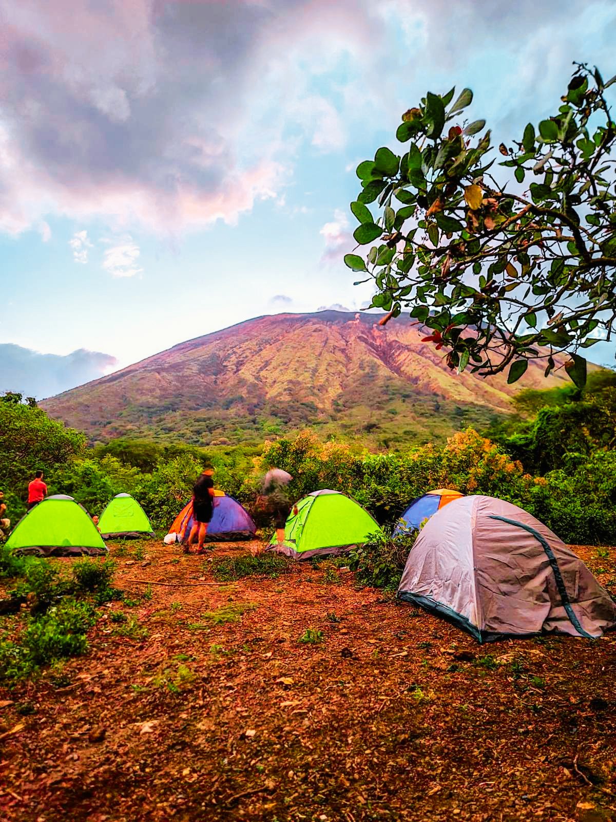 A group of tents set up in a clearing at the base of a large volcano, with hikers nearby, under a partly cloudy sky.