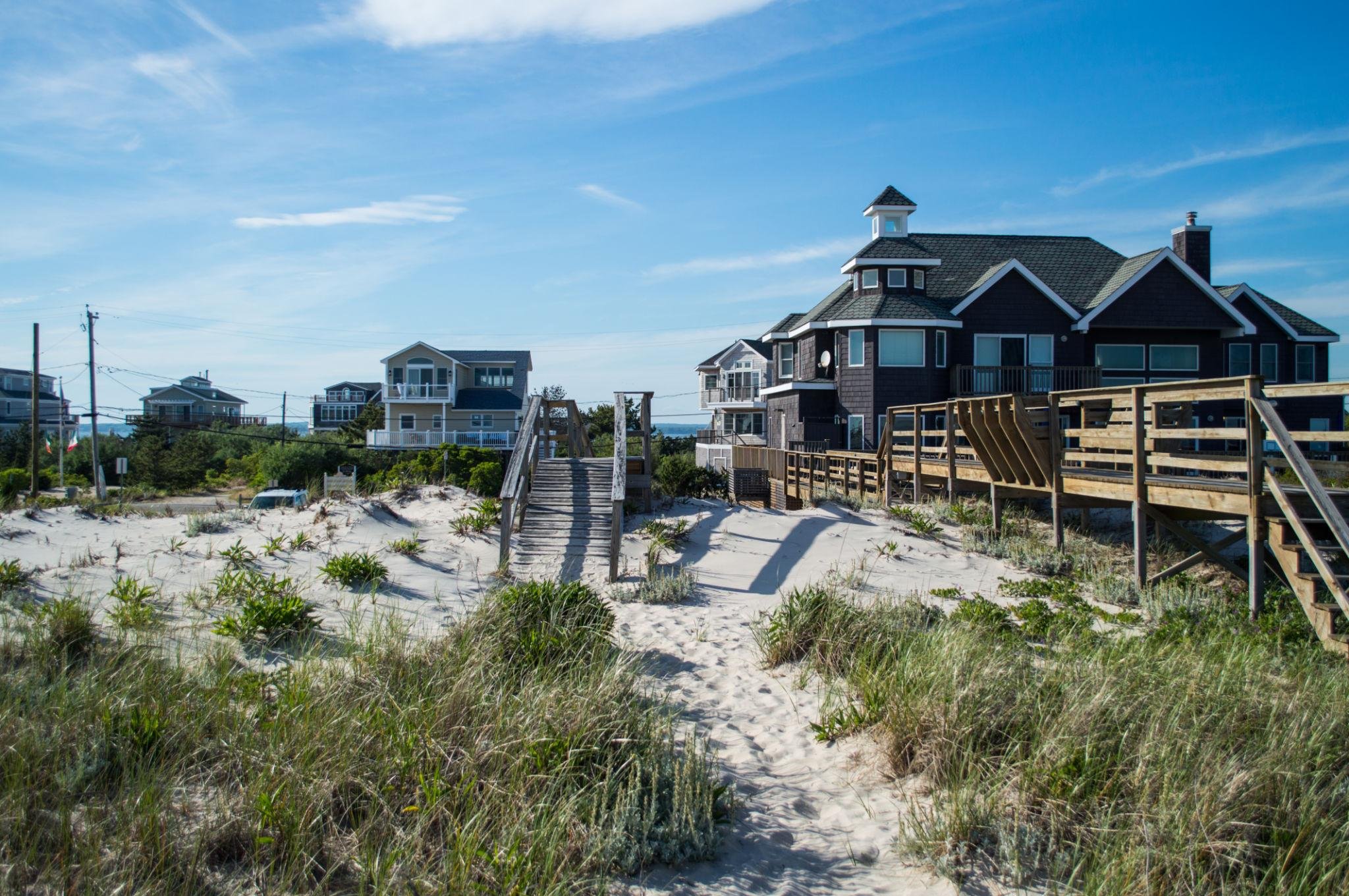 Beach houses with wooden walkways on sand dunes under a blue sky with scattered clouds.