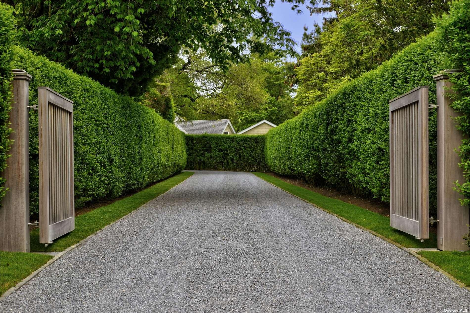 A gravel driveway with lush green bushes on both sides and tall trees overhead, leading to a house in the background with a pitched roof.