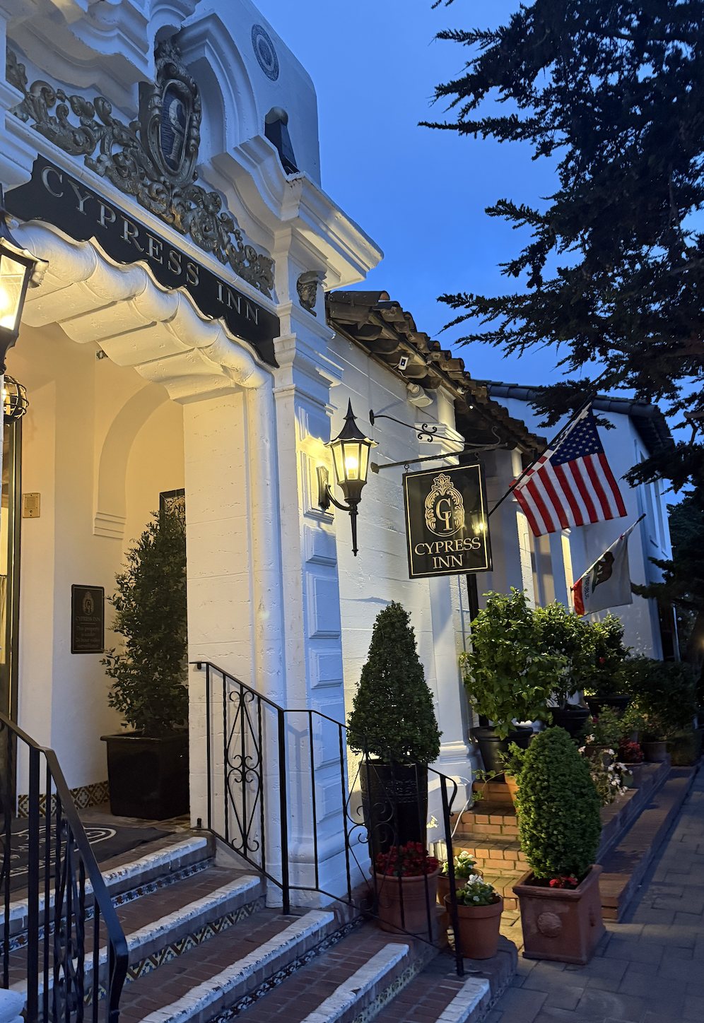 Exterior view of Cypress Inn at dusk, with colonial-style architecture, hanging lanterns, potted plants, and American flags.