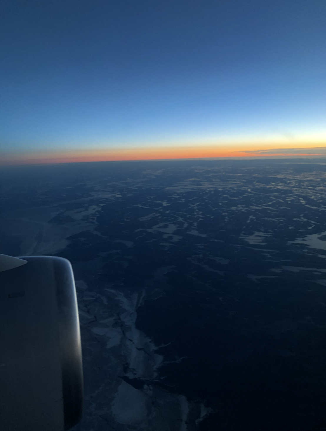 View of the sky at sunset from an airplane window, showing the horizon, clouds, and a portion of the airplane wing.