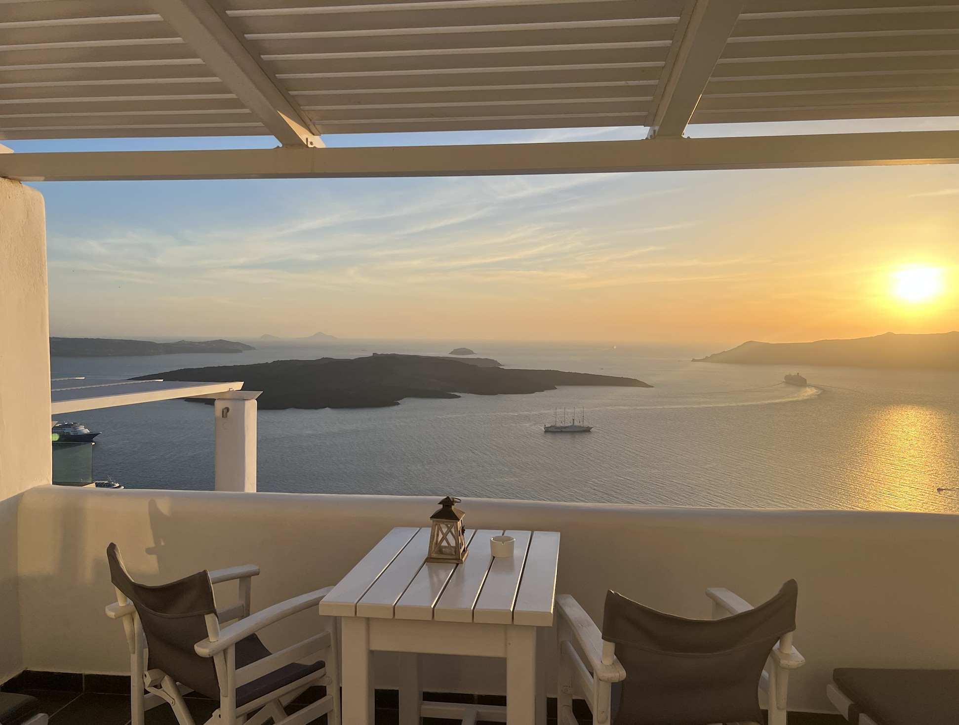 A balcony view during sunset overlooking the sea and islands, with a white table and chairs on the balcony, and a lantern and a cup on the table.