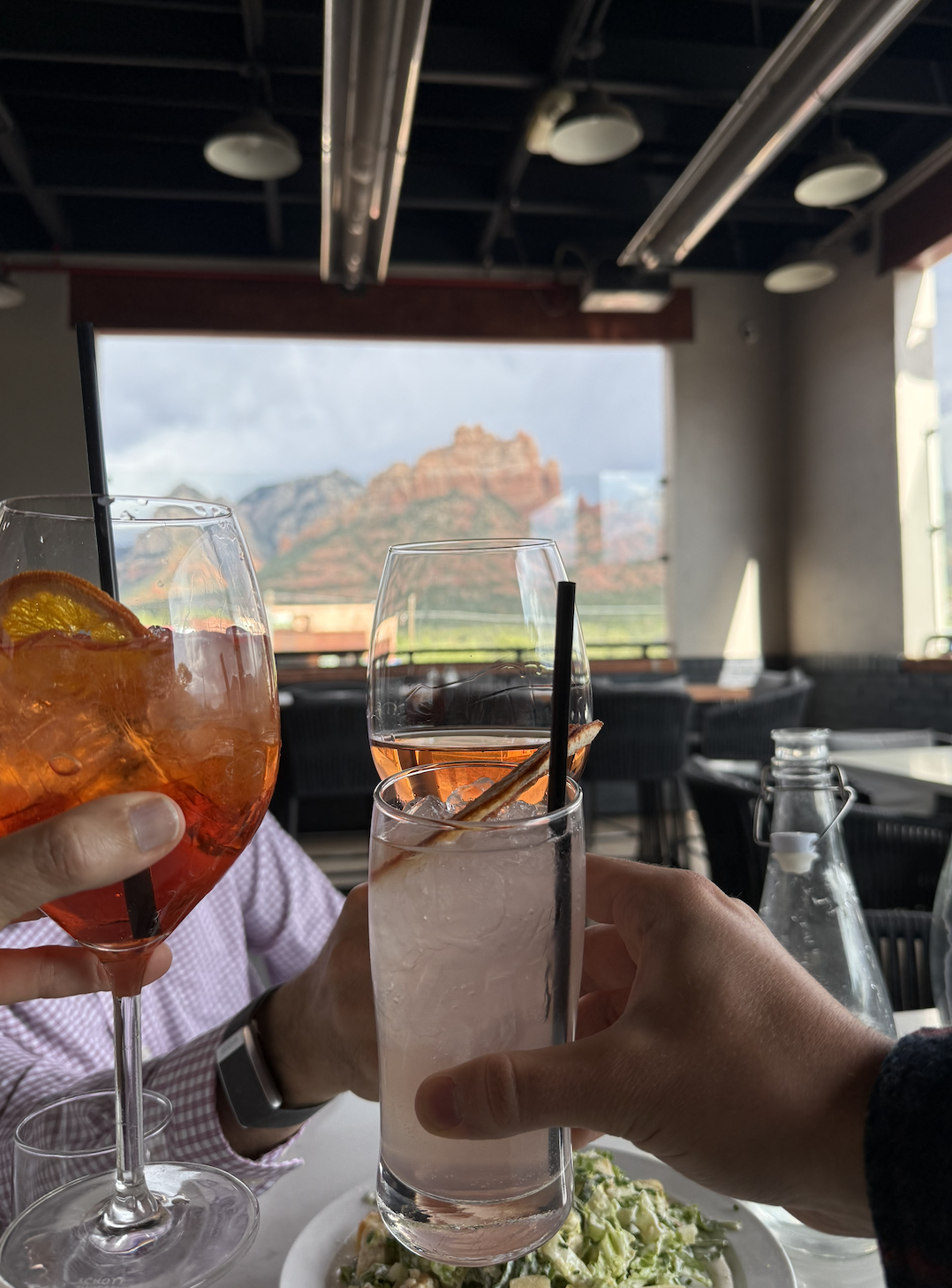 Three people toasting with colorful cocktails at a restaurant with a view of redrock mountains and blue sky.