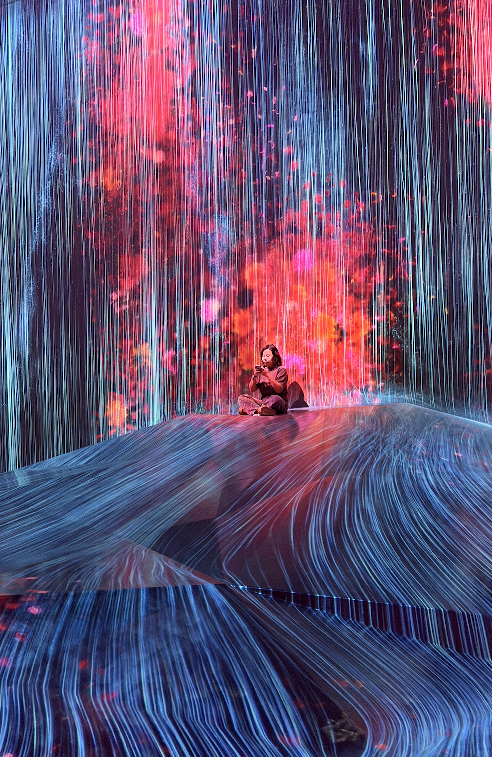 A person sitting cross-legged on a rock in front of a vibrant digital art installation with vertical light streams and colorful abstract patterns.