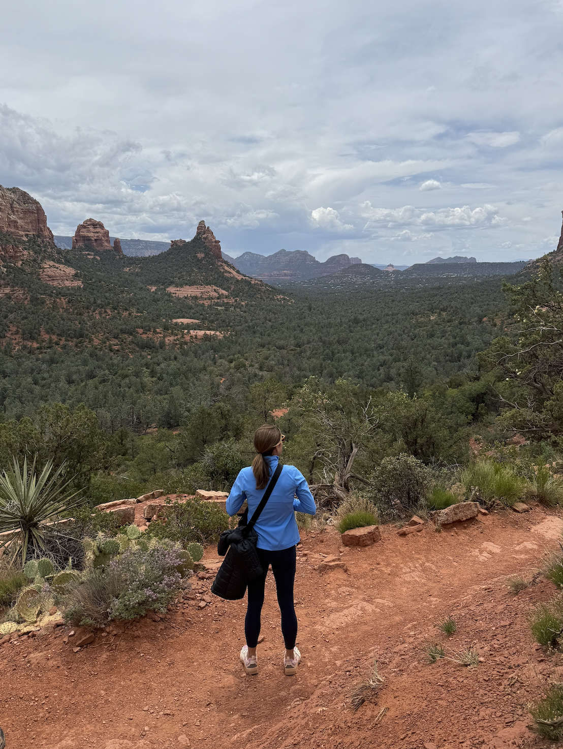 A woman in a blue jacket and black leggings hiking on a dirt trail in a desert landscape with red rocks, green vegetation, and a cloudy sky.