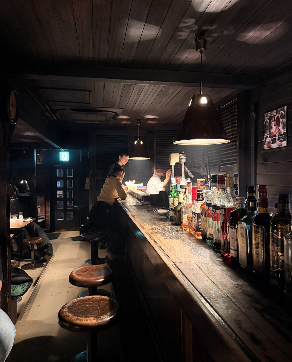 A dimly lit bar with a wooden counter, bottles of alcohol lined up, and patrons seated on stools. A bartender is serving a customer, with two hanging pendant lights overhead, and dark wood walls and ceiling.