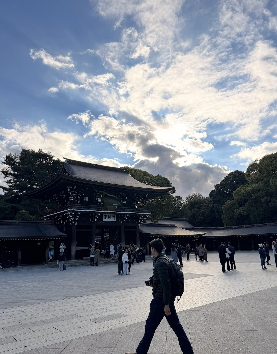 People visiting a traditional Japanese shrine with a wooden gate, surrounded by trees under a cloudy sky with the sun shining through.