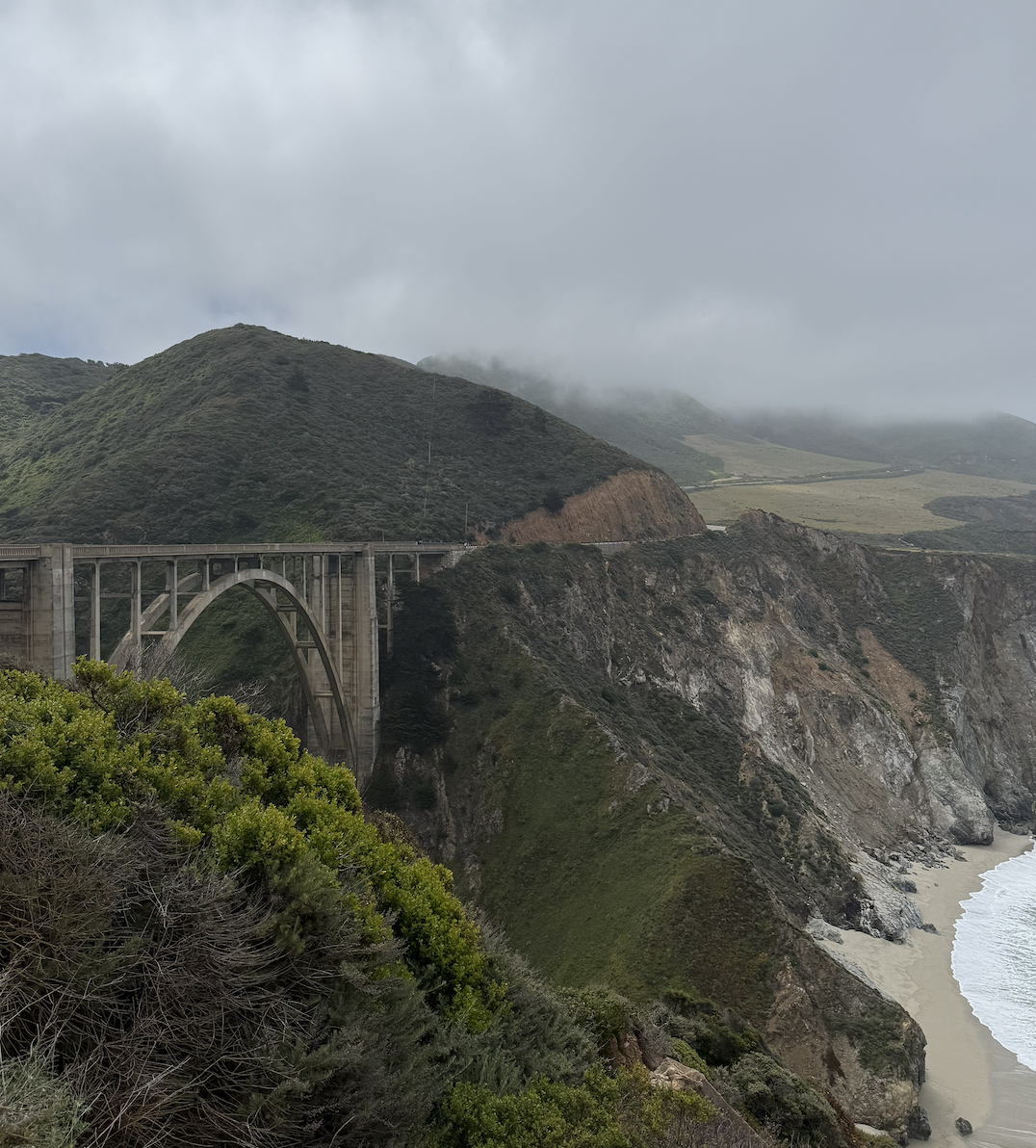 A scenic view of a coastal hillside with a concrete arch bridge extending across a deep valley, overlooking a beach and ocean under overcast skies.