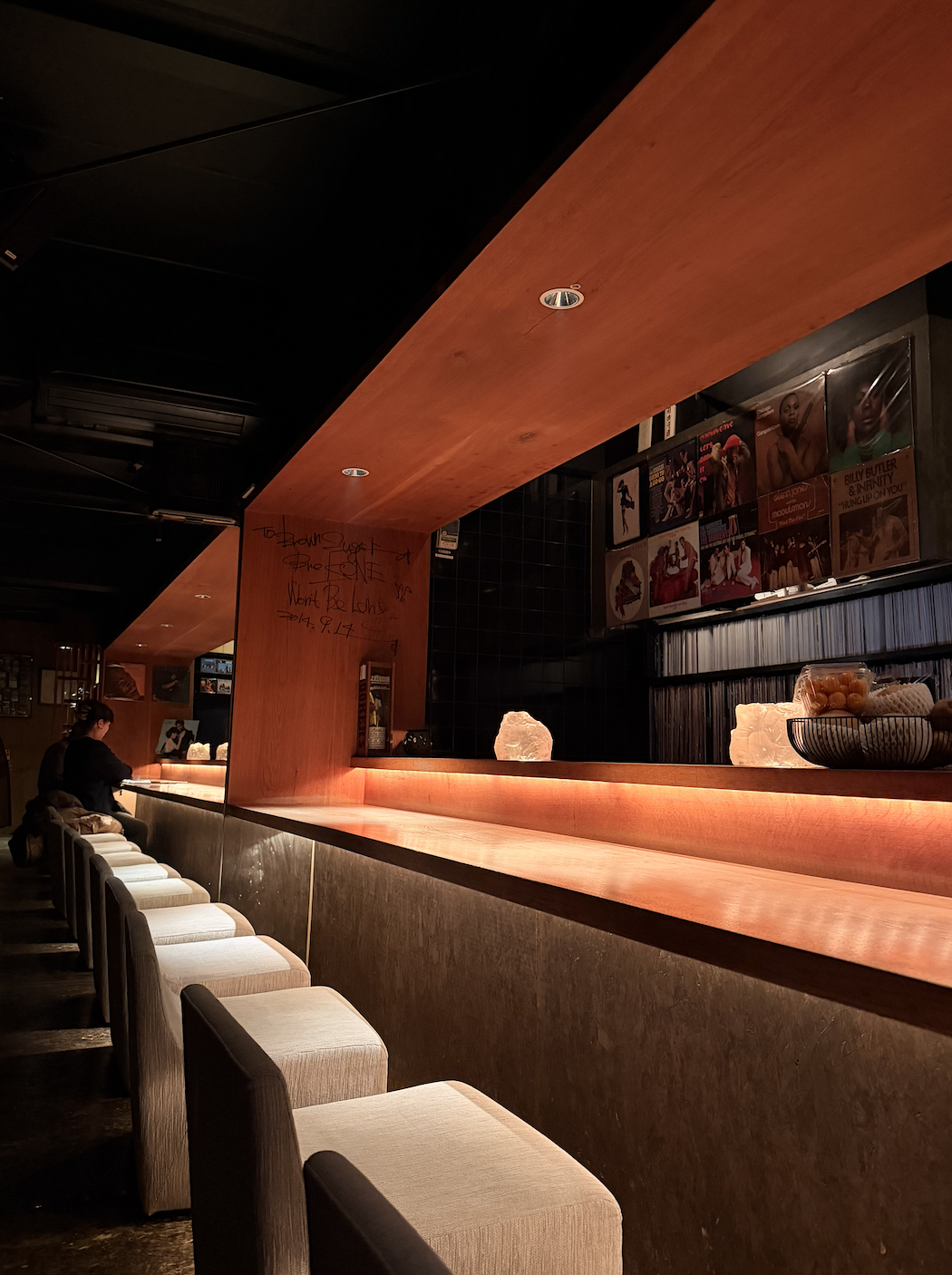 A dimly lit bar with a wooden counter, salt lamps, and framed photos or posters on the wall. There are chairs lined up along the counter, and a person is sitting at the bar.