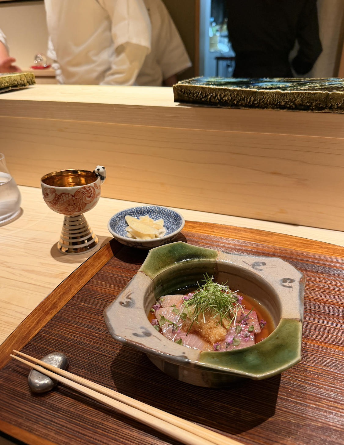 A Japanese dish with slices of raw fish garnished with greens, served in a decorative ceramic bowl on a wooden tray. There is a small dish of pickled ginger, a cup with a floral pattern, and a glass of water nearby. The restaurant setting has a woode