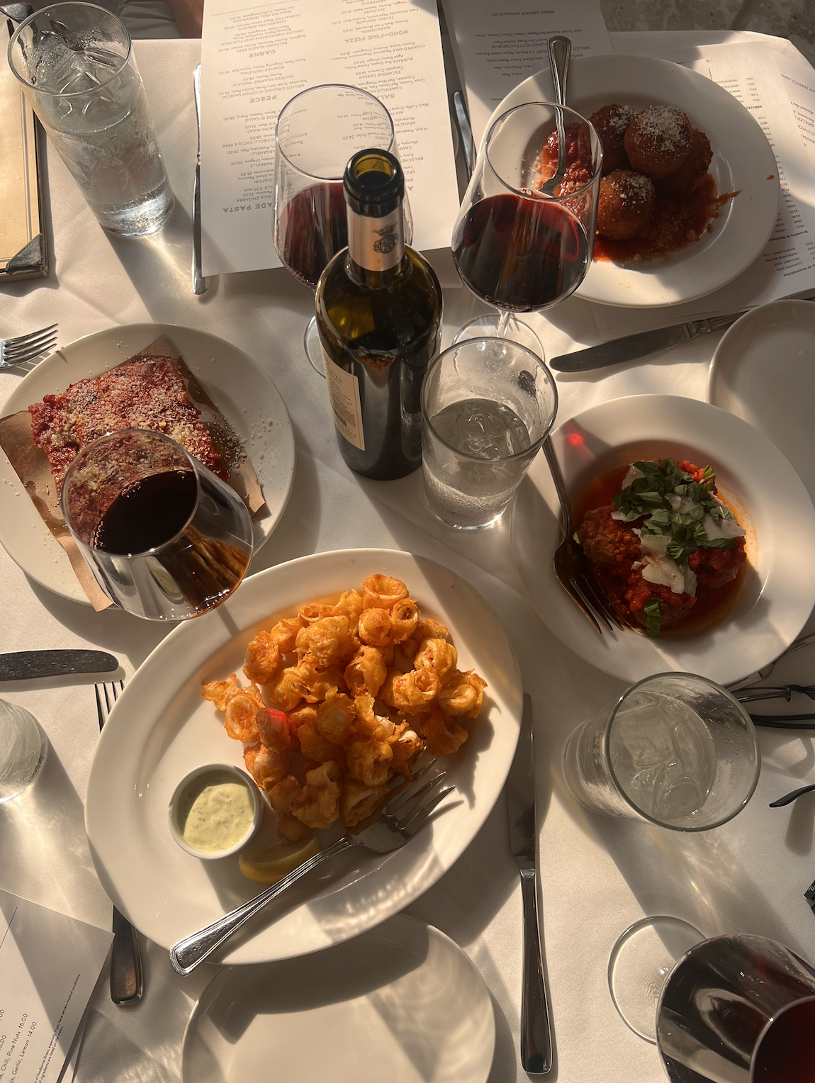 A table with various dishes including a plate of fried shrimp with dipping sauce, a meat lasagna, a plate of eggplant parmesan, and meatballs in tomato sauce with basil and cheese. There are several glasses of red wine, a bottle of wine, and glasses 