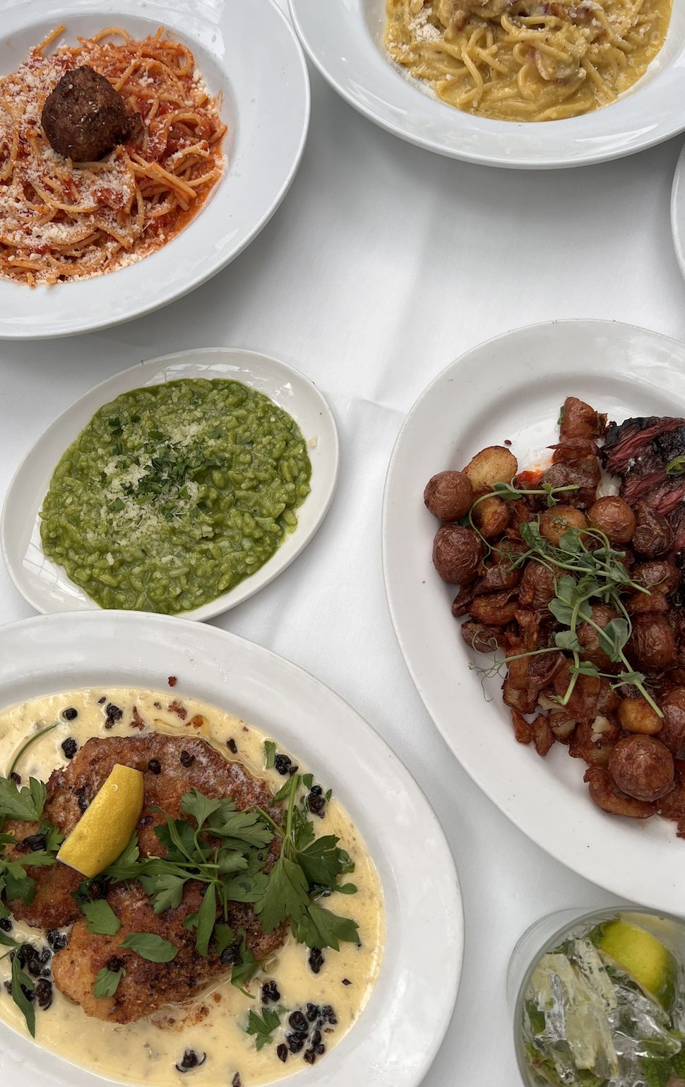 Various plates of food on a white table, including spaghetti and meatball, pasta with creamy sauce, calamari with herbs and lemon, mashed peas, braised beef with carrots and potatoes, and a glass of water with ice and lemon.