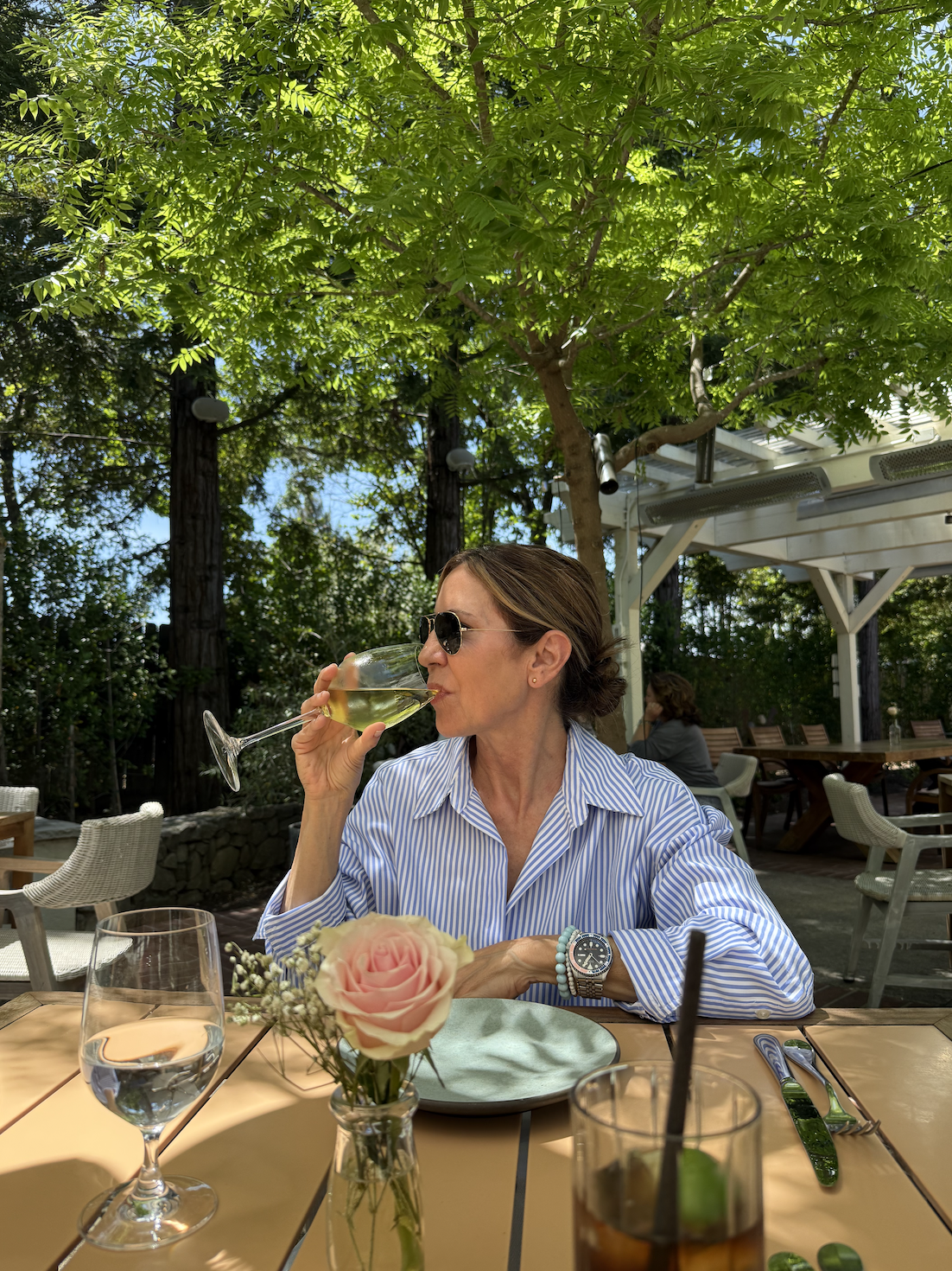 A woman wearing sunglasses and a striped blue shirt drinks white wine at an outdoor restaurant table surrounded by trees and greenery.