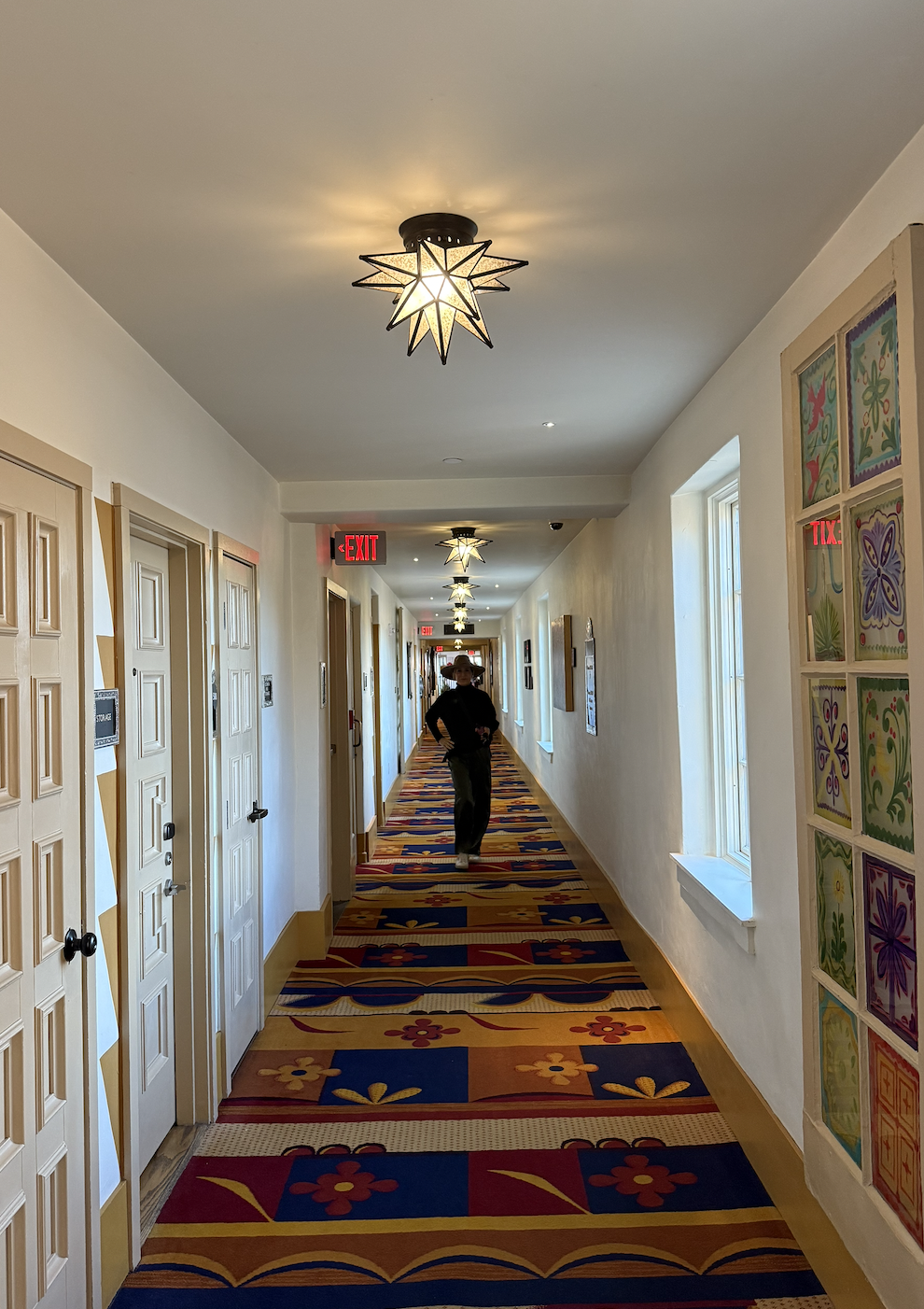 A hotel hallway with colorful floral-patterned carpet, white walls, wooden doors, and star-shaped ceiling lights. A person in black is walking towards the camera, with red exit signs visible.