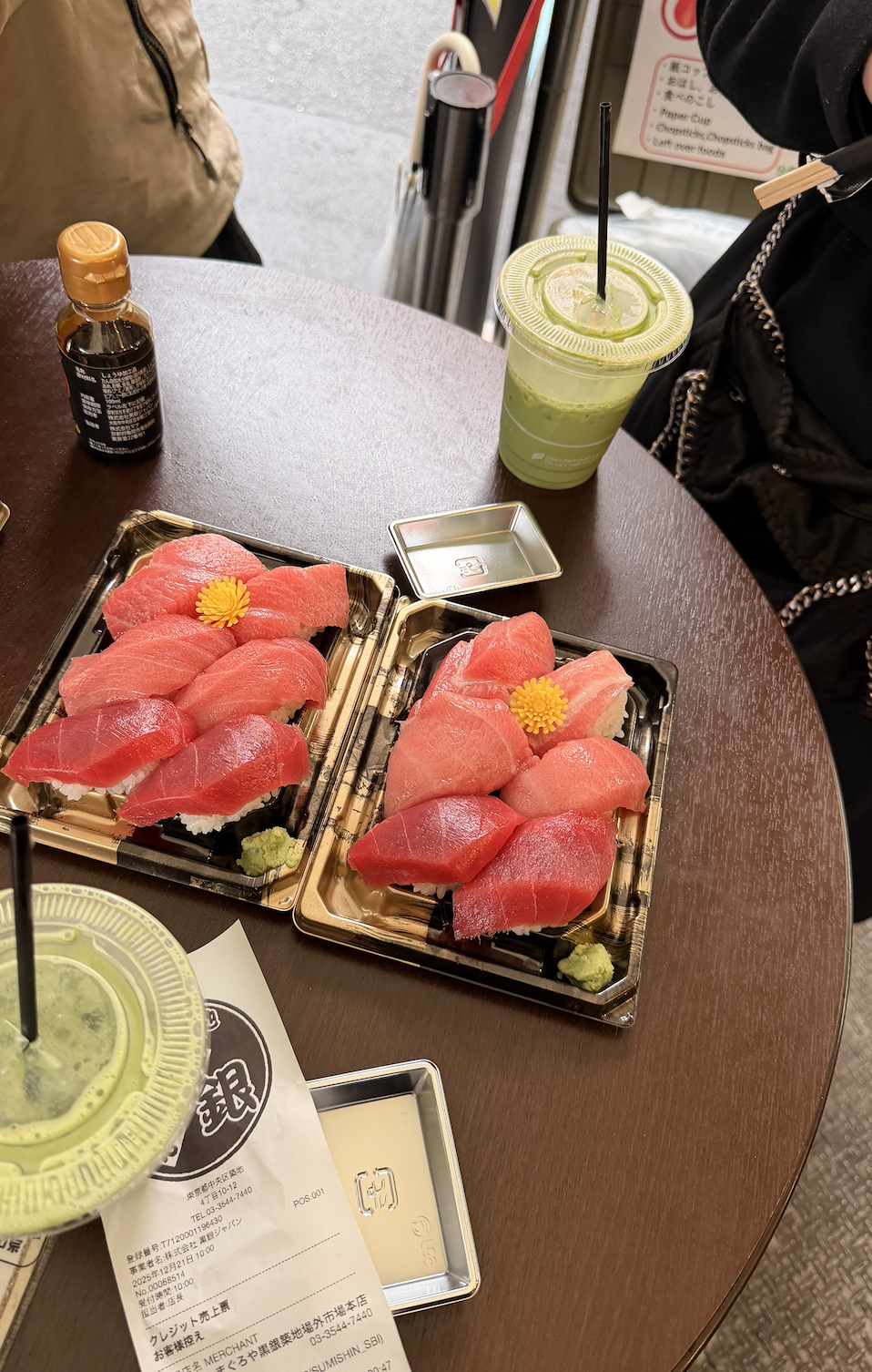 Two trays of assorted tuna sushi with wasabi and ginger, a green matcha iced drink with a black straw, a small sauce dish, a bottle of soy sauce, and a receipt on a wooden table at a restaurant.
