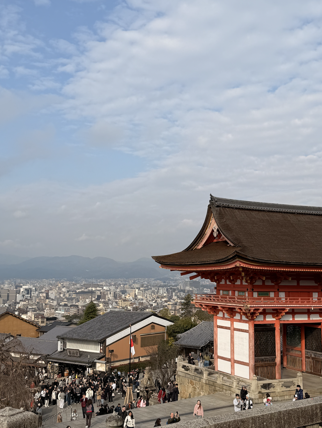 People gathered at a historic shrine in Kyoto with a red and white traditional building, overlooking a cityscape with mountains in the background on a cloudy day.