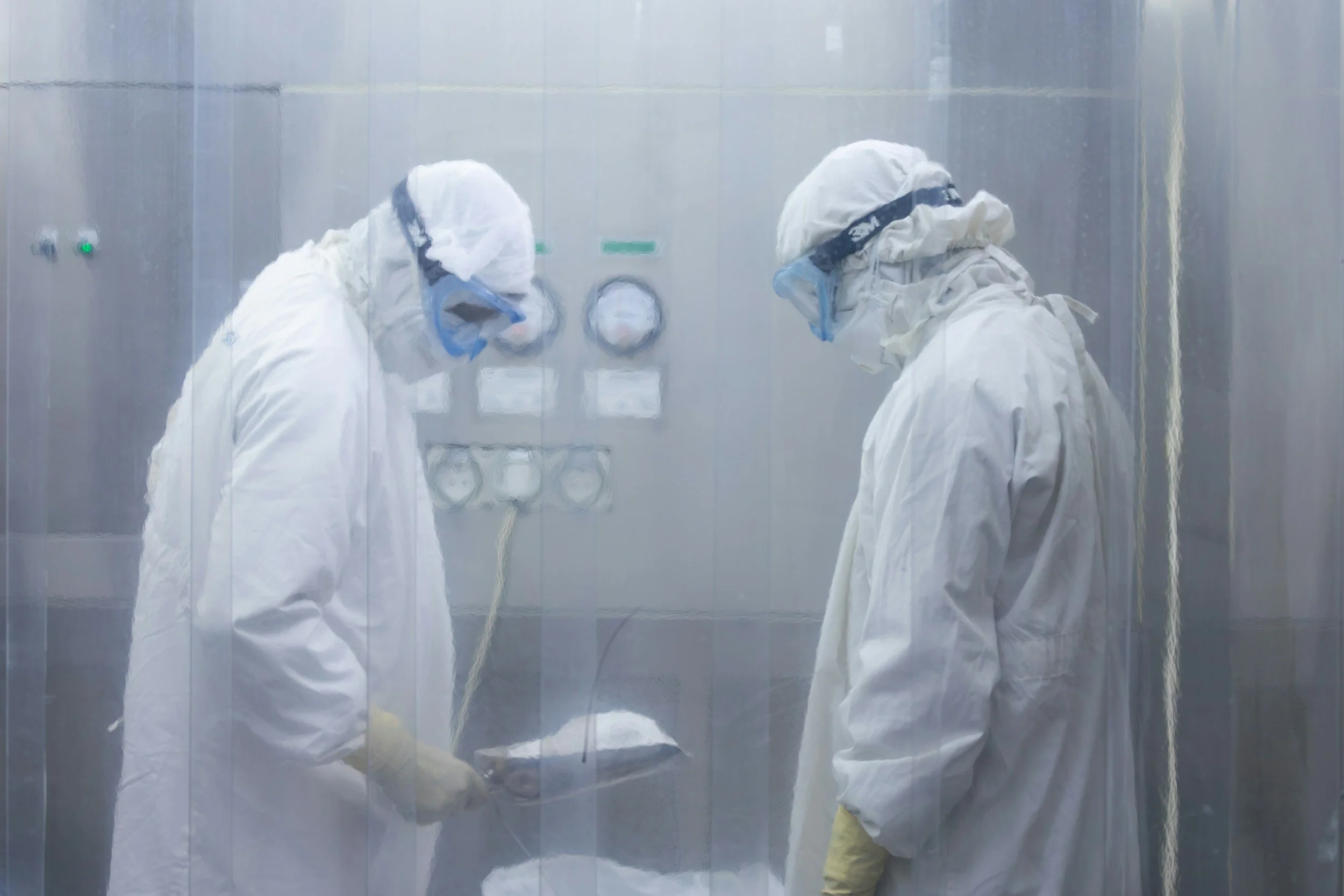 Two scientists or medical workers wearing full protective suits, masks, and face shields are working in a cleanroom or laboratory environment enclosed in a transparent barrier.
