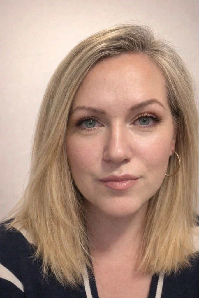 A woman with shoulder-length blonde hair, blue eyes, wearing hoop earrings and a dark top with light-colored accents.