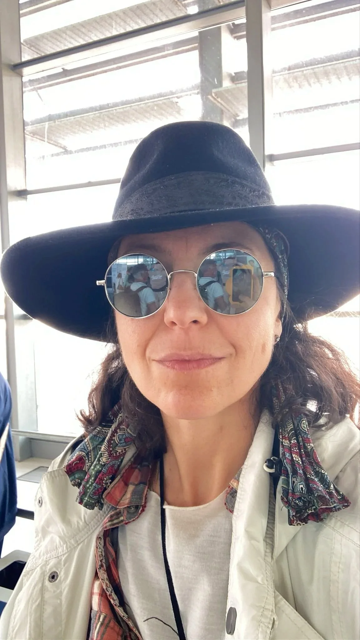 Gaye Gungor wearing a large black hat, round sunglasses, a white jacket, and a colorful scarf at an airport terminal.